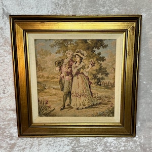 May include: A framed tapestry depicting two figures in historical attire walking through a landscape. The person on the left wears a coat and breeches, while the person on the right wears a long dress and hat. The frame is gold with a beige mat.