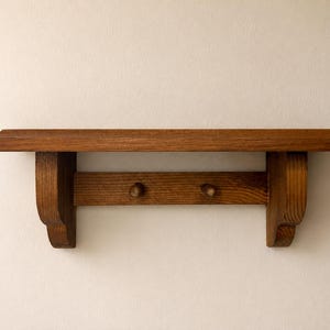 Pode incluir: Uma prateleira de parede de madeira castanho-média com uma superfície superior plana e borda chanfrada, suportada por dois suportes laterais curvos. Uma barra horizontal une os suportes, com dois pequenos ganchos de madeira arredondados.
