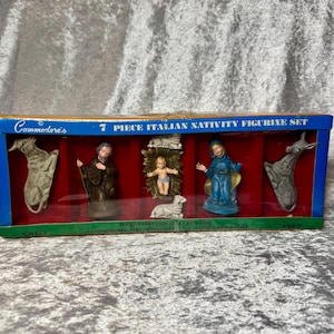May include: A 7-piece Italian nativity figurine set by Commodore in its original box. The set includes hand-painted figures of a kneeling man in brown robes, a woman in a blue robe, a baby in a gold manger, two sheep, a donkey, and a cow.