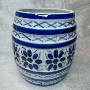 May include: A cylindrical ceramic vase with a white base and blue decorative elements. The vase features horizontal bands and floral patterns in blue. The design includes cross-hatch and floral motifs, creating a classic, decorative look.