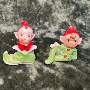 May include: Two ceramic elf figurines with pale skin, rosy cheeks, and pointed ears. One elf sits inside a light green shoe, while the other reclines on its side. Both figures wear bright red pointed hats and light green clothing accents.