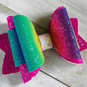 May include: A rainbow glitter hair bow with a white center. The bow is made of two layers of glitter fabric, with the top layer being a rainbow gradient and the bottom layer being a solid pink.