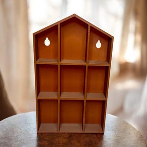 May include: A wooden, house-shaped display shelf with nine cubby holes. The shelf is a warm brown color and has two keyhole-shaped openings for hanging. The shelf is sitting on a round table.