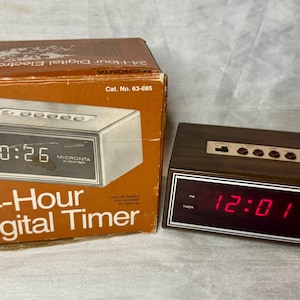 May include: A rectangular Micronta 24-Hour Digital Timer with a dark wood-grain finish and a red LED display showing 12:01. The device features top-mounted control buttons and is accompanied by its original brown cardboard packaging box.