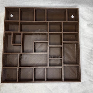 May include: A brown wooden wall-mounted display shelf with multiple compartments of varying sizes. The shelf has a square shape and includes two keyhole slots for hanging. The design is ideal for displaying small collectibles or decorative items.