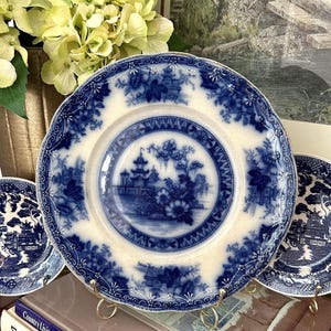 May include: A round ceramic plate featuring a blue and white transferware pattern of a pagoda, trees, and floral motifs. The rim has a scalloped edge with intricate floral detailing. The plate is displayed on a gold metal stand.