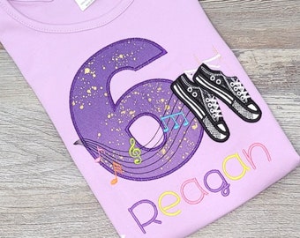 May include: Purple t-shirt with a sparkly purple number six, musical notes, and a pair of black sneakers. The name "Reagan" is written in colorful letters below the number.