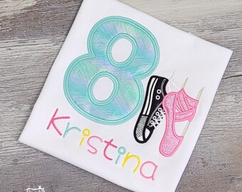 May include: A white t-shirt with a turquoise number eight, a black and white sneaker, and a pink ballet shoe. The name "Kristina" is embroidered in pink, yellow, and white.
