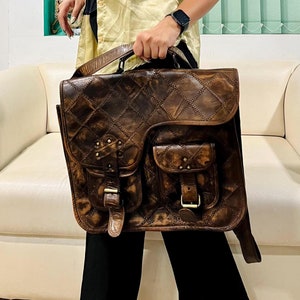 Puede incluir: Un bolso de cuero marrón con un diseño acolchado y varios bolsillos. El bolso tiene una correa larga y un asa superior.