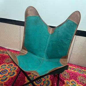 Pode incluir: Uma cadeira borboleta azul turquesa com detalhes em couro castanho e uma estrutura de metal preto. A cadeira está sentada sobre um tapete rosa e laranja com padrões.