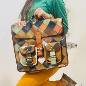 Puede incluir: Una mujer con una camisa verde y pantalones de color mostaza está sosteniendo una mochila grande de cuero con parches multicolores, con varios bolsillos y hebillas.