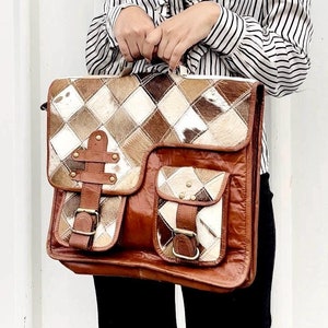 Puede incluir: Una mujer con una blusa a rayas blancas y negras sostiene un bolso de cuero marrón con un diseño de patchwork. El bolso tiene varios bolsillos y hebillas.
