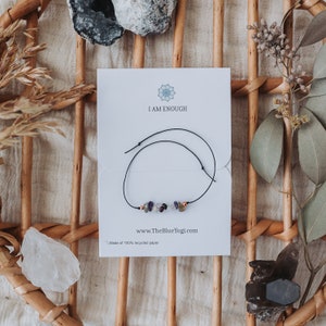 May include: A white card with the text "I AM ENOUGH" and a website address, "www.TheBlueYogl.com". A black string bracelet with small, colorful stones is displayed on the card. The card is made of 100% recycled paper.