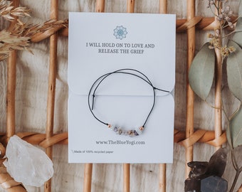 Könnte beinhalten: Ein schwarzes Armband aus Schnur mit einem kleinen Cluster aus hellgrauen und weißen Steinen und goldenen Akzenten. Das Armband wird auf einer weißen Karte mit dem Text "I WILL HOLD ON TO LOVE AND RELEASE THE GRIEF" und "www.TheBlueYogi.com" gezeigt.