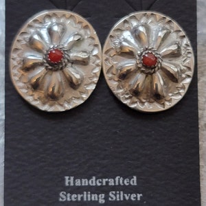 May include: A pair of handcrafted sterling silver earrings with a red coral stone in the center of each earring. The earrings are in the shape of a flower with a scalloped edge.