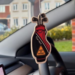 May include: A hanging car air freshener shaped like a red and black golf bag containing golf clubs. The bag features the text "Car-fect" and a yellow triangular sign with the black text "HOW'S MY DRIVING?" printed on the front surface.