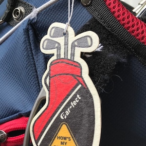 May include: A golf-themed car air freshener shaped like a golf bag with clubs. The air freshener is red, black, and white, with the text "Car-fect" and "How's my driving?" on a yellow triangle. It hangs from a golf bag.