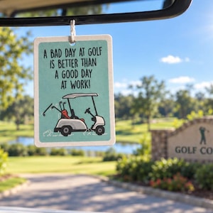 May include: A rectangular hanging air freshener with a light teal background, featuring a graphic of a white golf cart and the black text: A BAD DAY AT GOLF IS BETTER THAN A GOOD DAY AT WORK! The item is suspended by a white string from a rearview mirror.