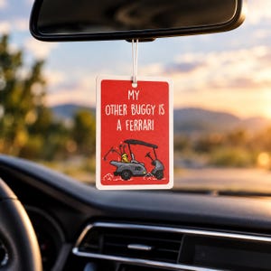 May include: A rectangular car air freshener hanging from a rearview mirror. The red air freshener features a graphic of a grey golf cart with the text MY OTHER BUGGY IS A FERRARI printed in white letters above the illustration of the vehicle.