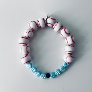 May include: A blue and white beaded bracelet with the word "BREW CREW" spelled out in white letters on the beads. The bracelet is made with baseball-themed beads.