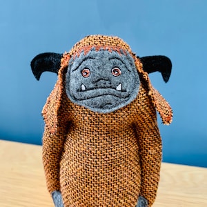 May include: A brown and orange plush toy with a gray felt face and embroidered eyes and teeth. The toy has a shaggy, furry texture and large, floppy ears.