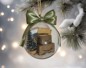 May include: A clear glass Christmas ornament with a green satin bow and ribbon. Inside the ornament are miniature cardboard boxes with the Amazon logo, a small Christmas tree, and artificial snow.
