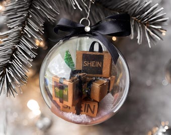 May include: A clear glass Christmas ornament with a black ribbon and bow. Inside the ornament are miniature cardboard boxes with the word "SHEIN" printed on them. The boxes are surrounded by white faux snow.