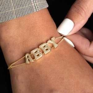 Peut inclure: Un bracelet en chaîne dorée avec le nom "ABBY" écrit en lettres majuscules. Chaque lettre est incrustée de petites pierres de zircone cubique claires.
