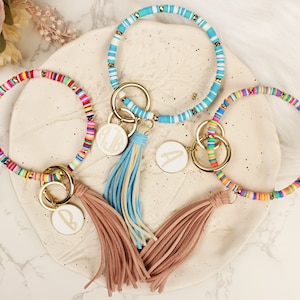 May include: Three colorful beaded keychains with gold key rings and tassels. Each keychain has a white round charm with a gold letter on it. The charms read "A", "B", and "LIB".