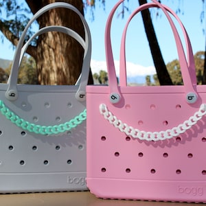 May include: Two tote bags, one gray and one pink, with matching handles and decorative chain accents. The bags have a series of small holes and the brand name "bogg" is visible on the bottom. The bags are outdoors with a blurred background.