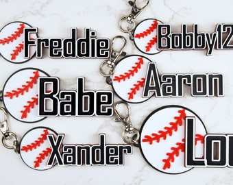May include: Six baseball-themed keyrings with different names on them. The keyrings are white with red stitching and black lettering. The names on the keyrings are Freddie, Babe, Aaron, Bobby123, Lou, and Xander.