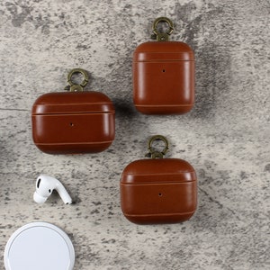 May include: Three brown leather protective cases for wireless earbuds, each featuring a bronze-toned metal ring attachment at the top. A single white wireless earbud and a white circular charging pad are positioned near the cases.