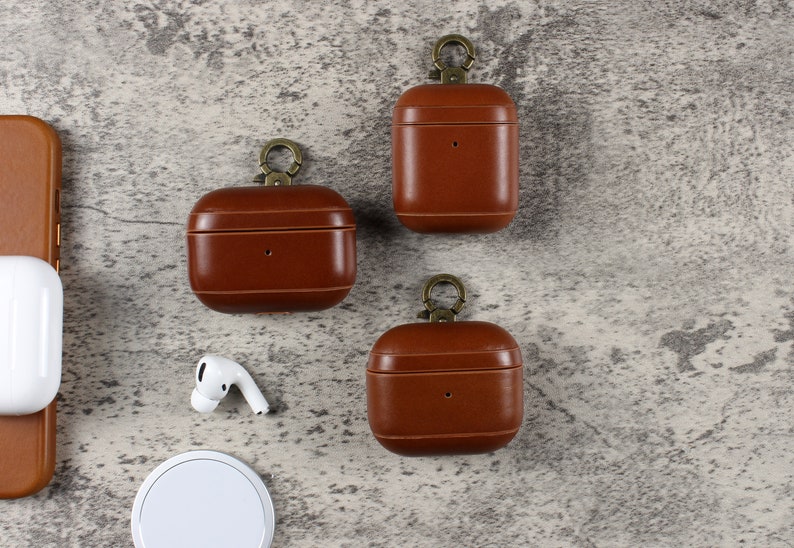 May include: Three brown leather protective cases for wireless earbuds, each featuring a bronze-toned metal ring attachment at the top. A single white wireless earbud and a white circular charging pad are positioned near the cases.