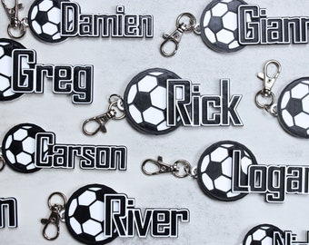 以下が含まれることがあります： 名前入りの白黒のサッカーボールキーホルダーが10個。名前は、ダミアン、グレッグ、リック、カーソン、ローガン、リバー、ジャンニ、エヴァン、そして読めない名前が2つです。