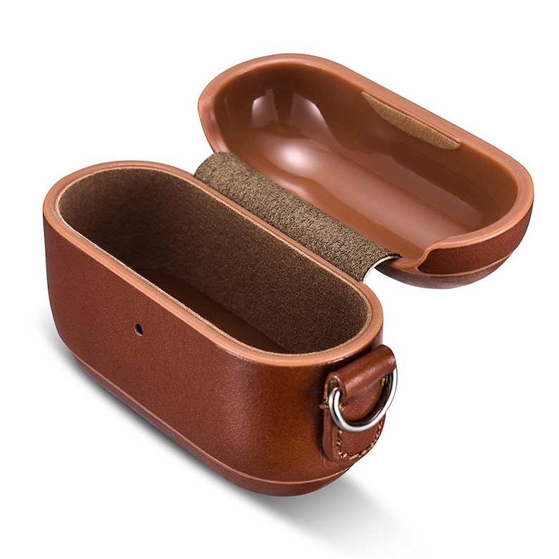 May include: An open brown protective case for wireless earbuds with a smooth exterior finish, a textured interior lining, a hinged lid, and a small metal D-ring attached to the side for a strap. The case features a small circular cutout on the front.