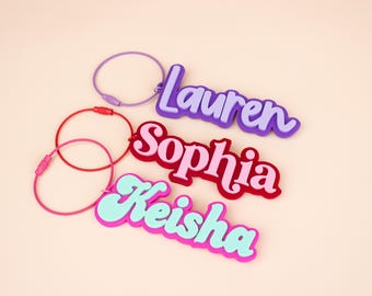 May include: Three custom name keychains featuring the names Lauren, Sophia, and Keisha in layered, colorful acrylic script. Each keychain is attached to a thin, circular metal wire loop in matching purple, red, and pink colors respectively.