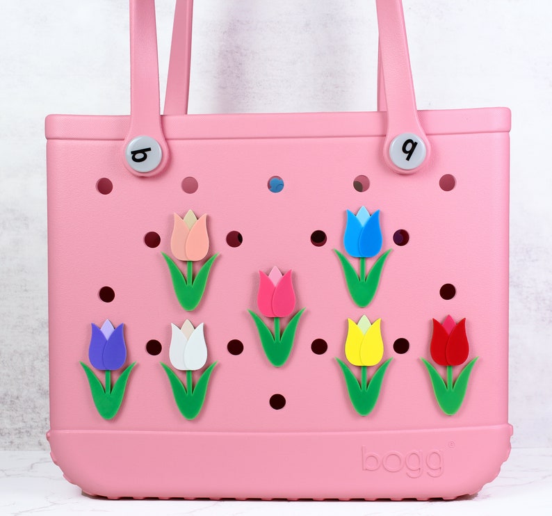 May include: Pink  Boga bag with a variety of colorful tulip charms attached to the front. The bag has a white label with the word "boga" printed on it.