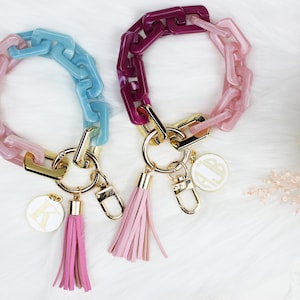 May include: Two colorful chain keychains with gold accents and tassels. One keychain is pink and blue, the other is pink and purple. Each keychain has a gold charm with a monogram.