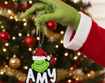 May include: A Christmas ornament featuring the Grinch wearing a Santa hat, with the name "AMY" below. The ornament is held by a green, furry hand with long fingers, against a backdrop of a decorated Christmas tree with red and gold ornaments.