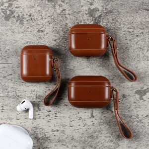 May include: Three brown leather protective cases for wireless earbuds, each featuring a matching leather wrist strap attached with a metal rivet. A brown leather phone case, a white wireless earbud, and a white charging case are also visible.