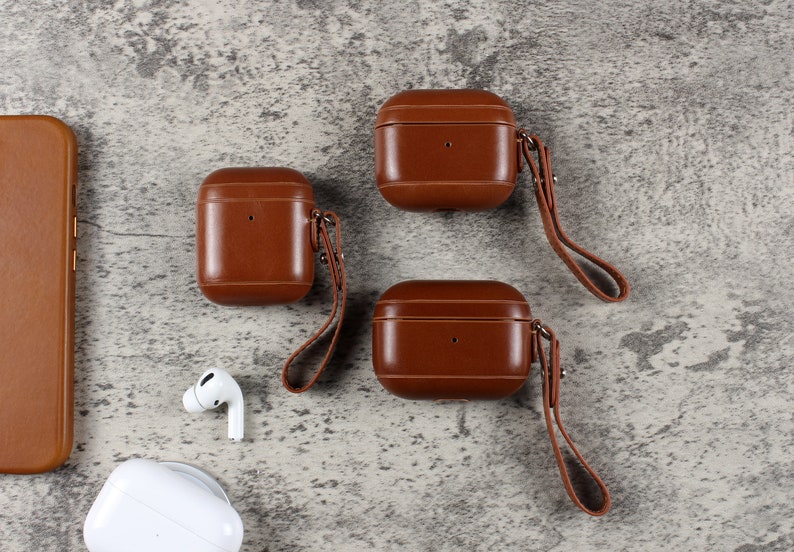 May include: Three brown leather protective cases for wireless earbuds, each featuring a matching leather wrist strap attached with a metal rivet. A brown leather phone case, a white wireless earbud, and a white charging case are also visible.