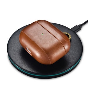 May include: A brown leather protective case for wireless earbuds, featuring a small circular cutout for a metal ring attachment and a visible green LED indicator light, resting on a circular black wireless charging pad with a thin teal accent.