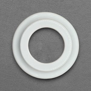 Puede incluir: Un objeto circular blanco con un diseño en forma de anillo. El objeto tiene un acabado liso y brillante y está compuesto por dos círculos concéntricos, creando una abertura central. El objeto está sobre un fondo gris.