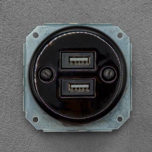 Pode incluir: Uma tomada de parede preta circular com duas portas USB empilhadas centradas numa placa frontal brilhante. A unidade está montada numa estrutura metálica quadrada com quatro orifícios para parafusos nos cantos, com um design funcional.