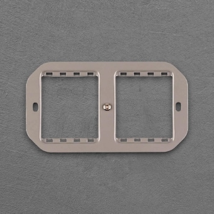 Può includere: Copertura per presa elettrica color argento con due aperture quadrate. La copertura ha una vite centrale e piccole fessure rettangolari lungo i bordi superiore e inferiore. Due piccoli fori sono sui lati.