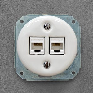 Op de afbeelding: Een witte wandplaat met dubbele poort en twee Cat5e Ethernet-aansluitingen. De ronde afdekplaat is bevestigd met twee zilverkleurige platte schroeven en gemonteerd op een vierkant metalen frame met zichtbare montagegaten in elke hoek.