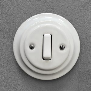 Può includere: Un interruttore della luce in ceramica bianca con un interruttore rettangolare e due piccole viti. L'interruttore è montato su una parete grigia strutturata. L'interruttore ha un design classico e vintage.