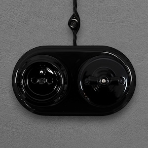 May include: A glossy black oval wall plate featuring a single electrical socket on the left and a rotary light switch on the right. The unit is connected to a black braided fabric cable that extends upwards from the centre of the top edge.