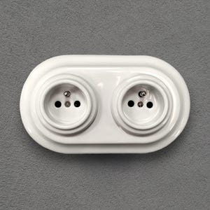 Może przedstawiać: Białe, podwójne gniazdo elektryczne z dwoma okrągłymi gniazdami. Gniazdo jest zamontowane na szarej ścianie. Każde gniazdo ma trzy małe otwory na bolce wtyczki.