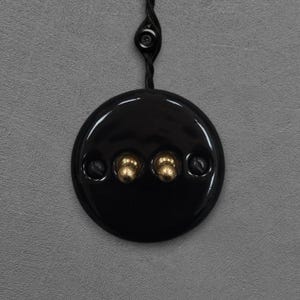 Pode incluir: Um interruptor de luz preto e redondo com dois botões dourados e dois parafusos. O interruptor está ligado a um cabo preto com um pequeno botão redondo. O fundo é de cor cinza lisa.